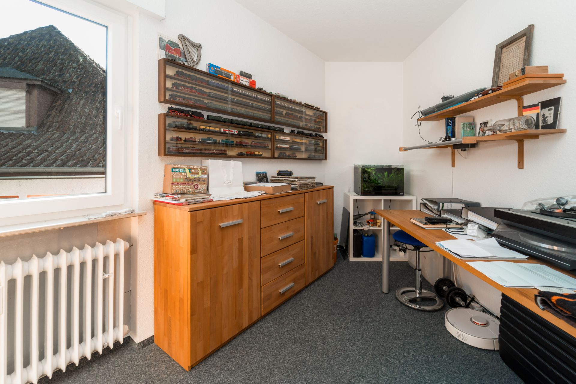 Kleines Arbeitszimmer mit Holzschrank, Schreibtisch, Regalen und Modellautos an der Wand.