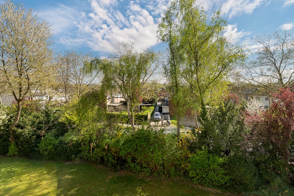 Blick auf grünen Garten mit Bäumen und Sträuchern, dahinter Wohnhäuser und blauer Himmel.