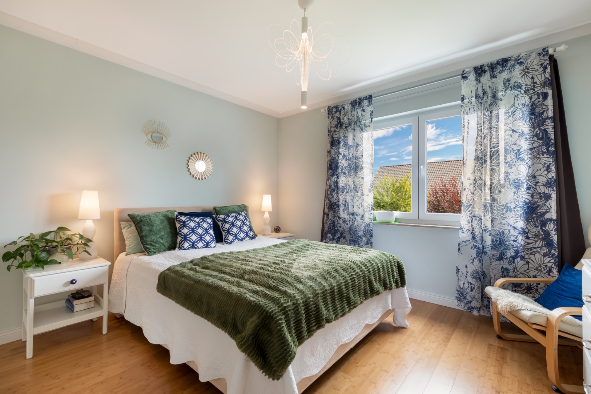 Helles Schlafzimmer mit Doppelbett, Nachttischen, Lampen und großem Fenster mit gemusterten Vorhängen.