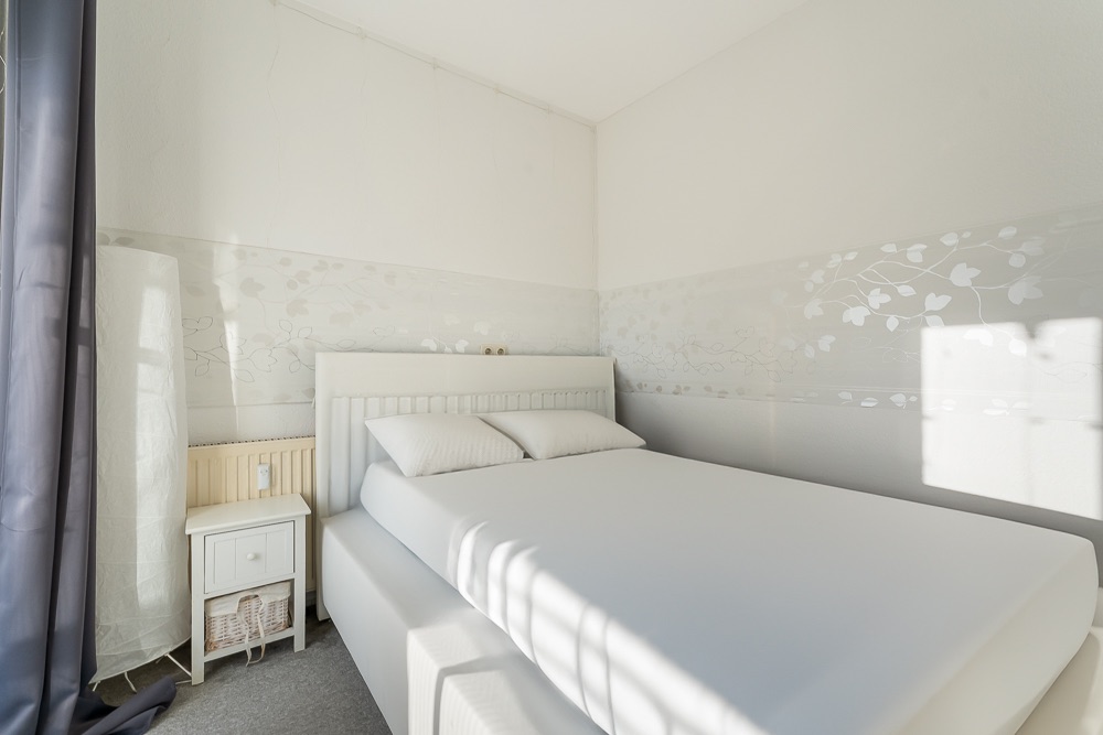 Helles Schlafzimmer mit Doppelbett, weißer Bettwäsche und Nachttisch neben einem Fenster.