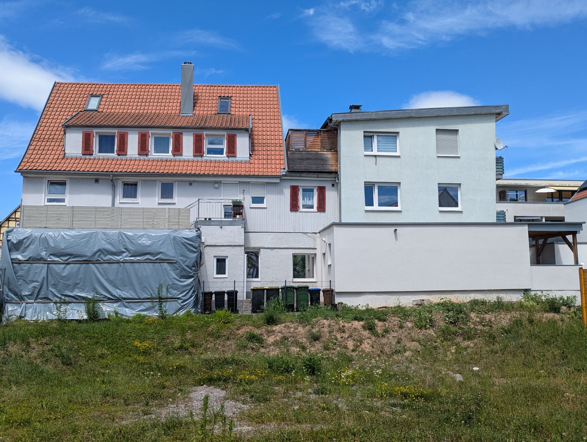 Mehrstöckiges Wohnhaus mit rotem Dach, weißen Wänden und roten Fensterläden, davor Wiese.