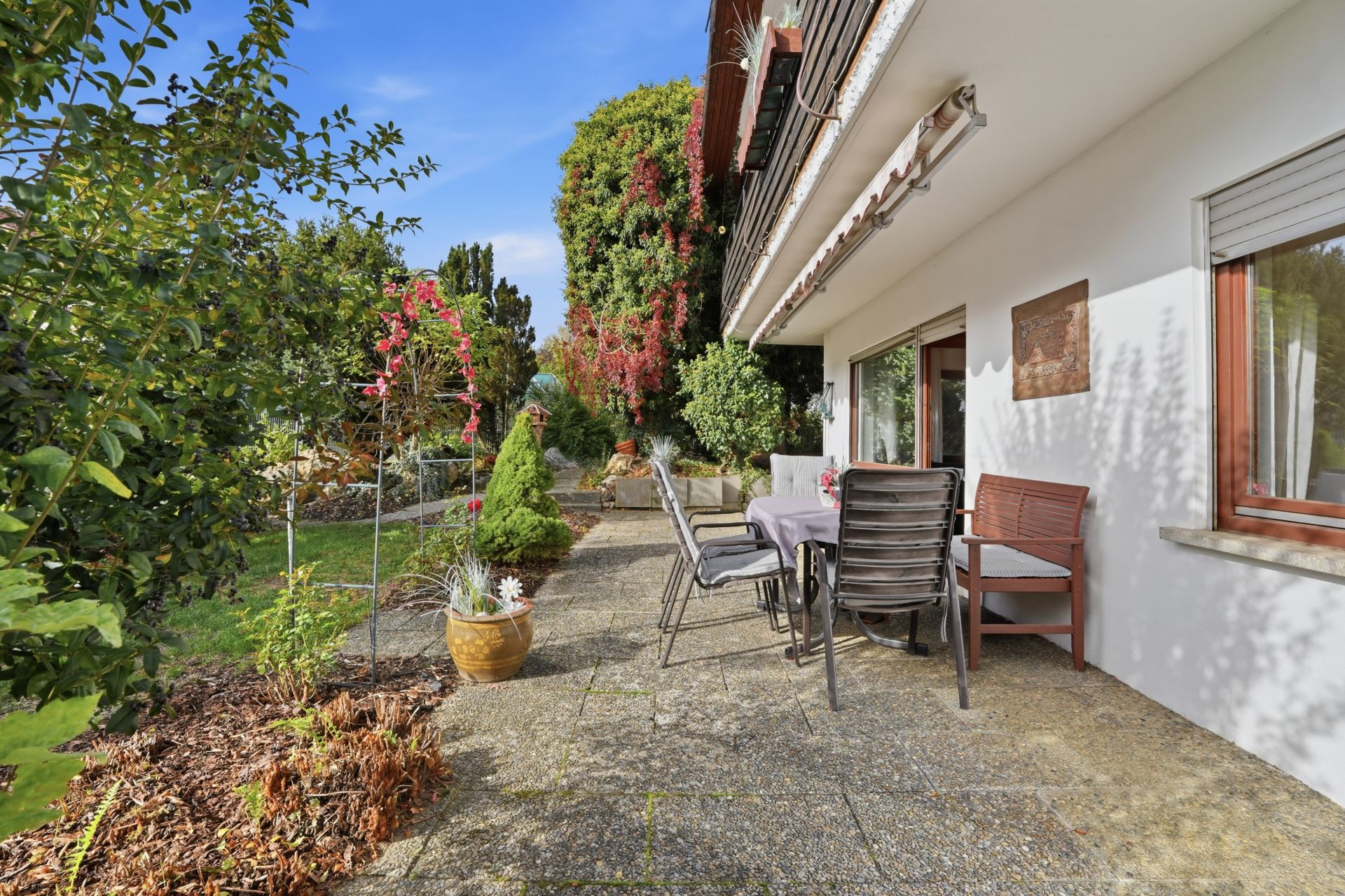 Terrasse mit Tisch, Stühlen und Pflanzen, neben einem Garten und einem Wohnhaus.