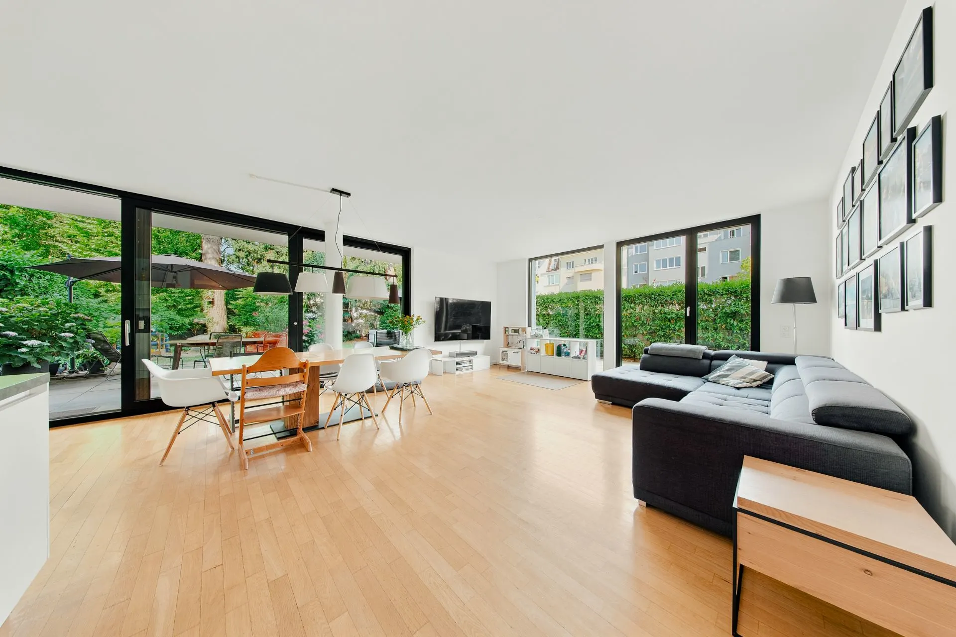 Helles Wohnzimmer mit Holzboden, großer Couch, Esstisch und großen Fenstern zum Garten.