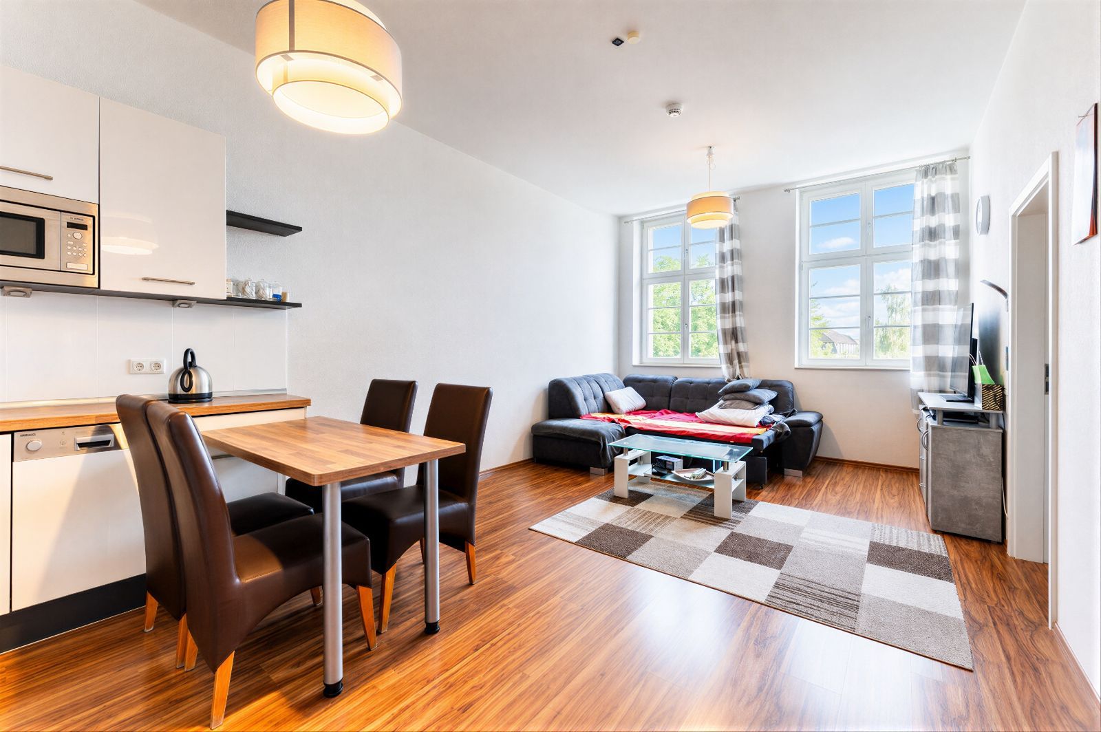 Helles Wohnzimmer mit Esstisch, Küche, Sofa, Teppich und großen Fenstern.