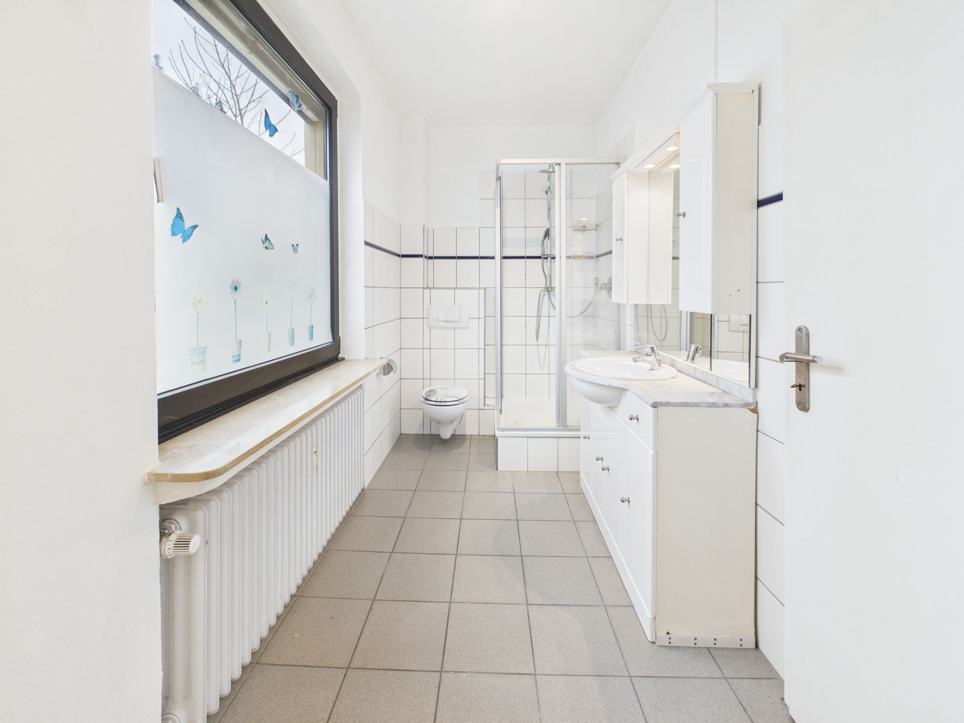 Helles Bad mit großer Fenster, Dusche, Toilette und weißem Waschbecken mit Schränken.