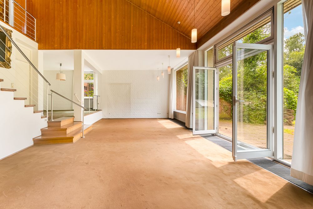 Helles Wohnzimmer mit großer Fensterfront, offener Tür, Teppichboden und Holzdecke.