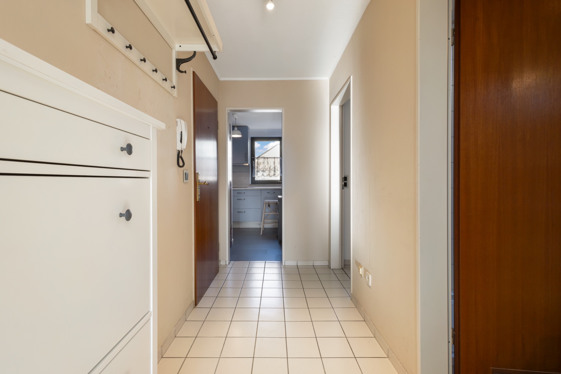 Heller Flur mit Fliesenboden, Schuhschrank, Holztüren und Blick in einen weiteren Raum mit Fenster.