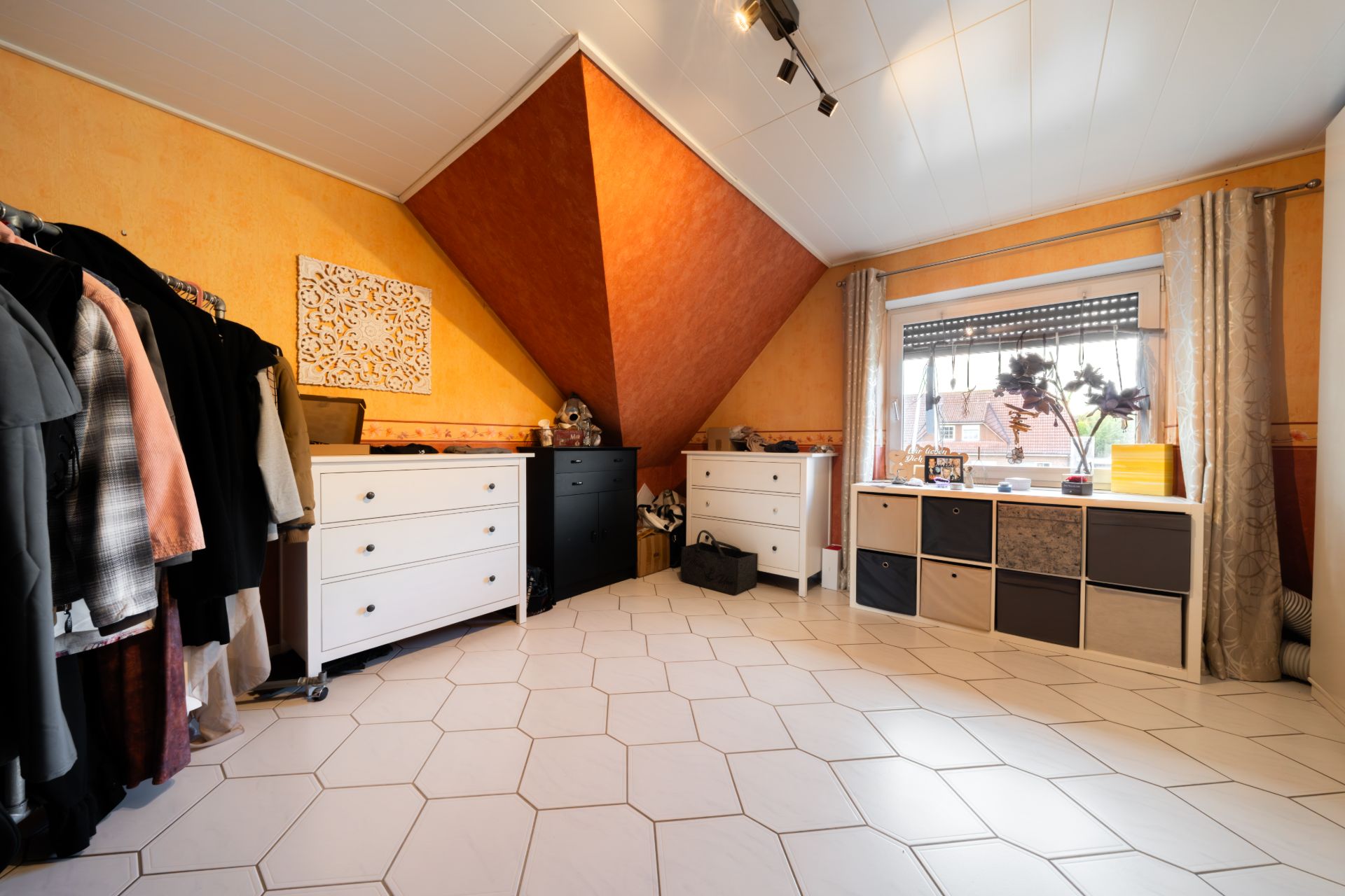 Zimmer mit weißen Kommoden, Kleiderständer und einem Fenster, hell gefliest und mit schräge Decke.