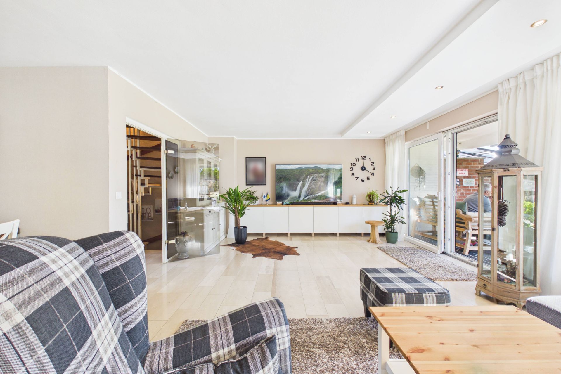 Helles Wohnzimmer mit karierten Sesseln, Holztisch, Pflanzen und großen Fenstern zur Terrasse.