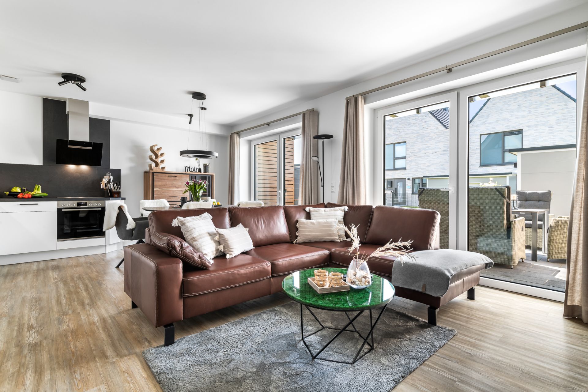 Helles Wohnzimmer mit braunem Sofa, Glastisch und offener Küche, große Fenster mit Blick nach draußen.