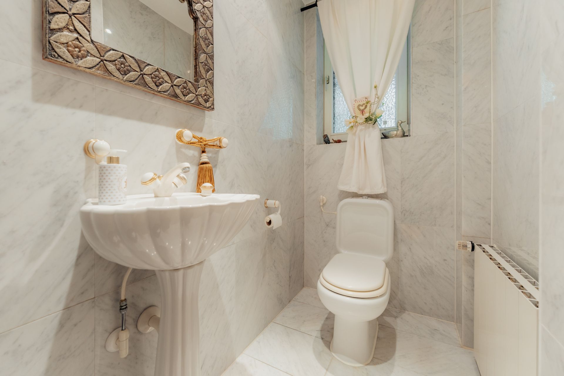 Kleines Badezimmer mit weißer Toilette, Waschbecken, Spiegel und Fenster mit Vorhang.