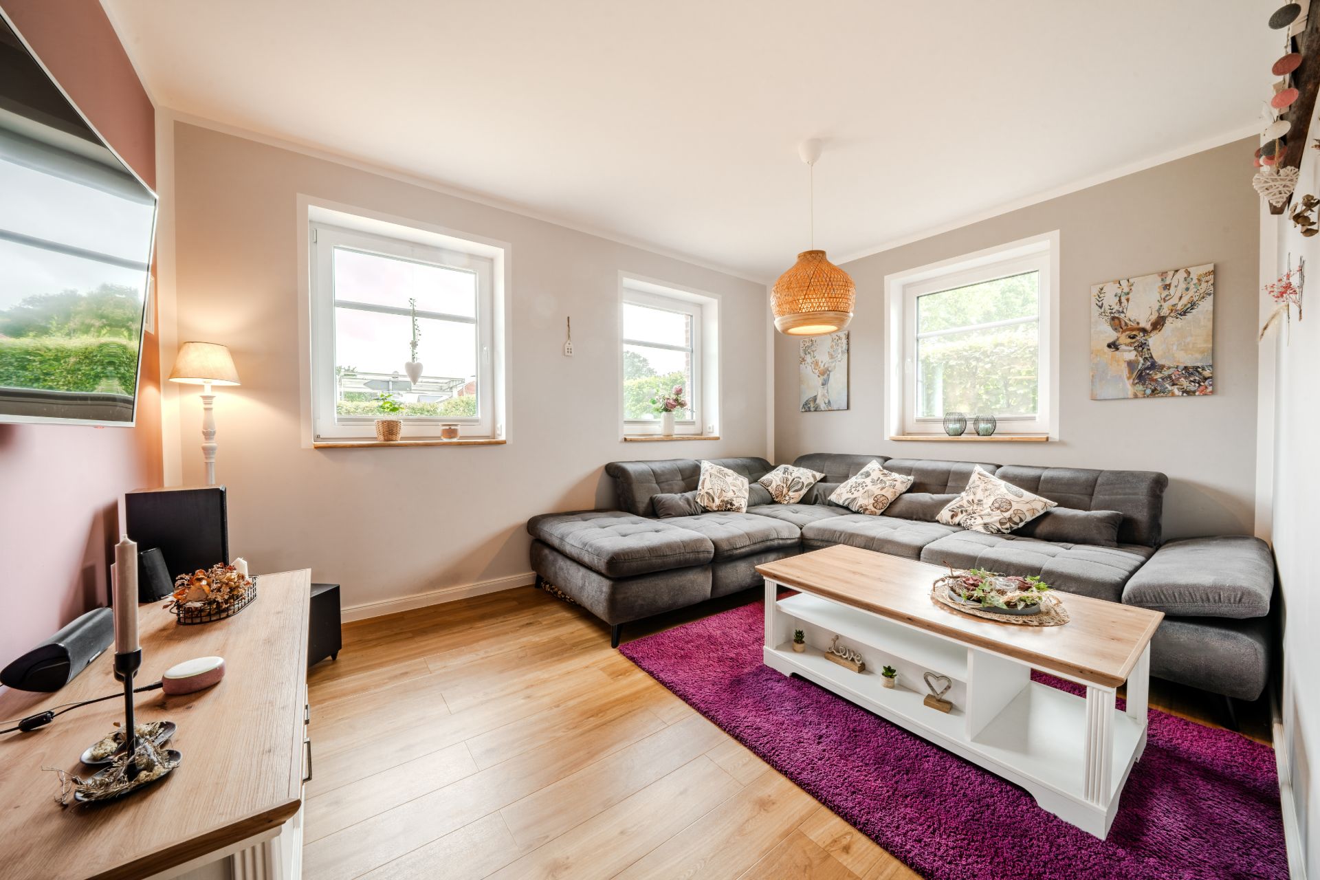 Helles Wohnzimmer mit grauem Ecksofa, Couchtisch, lila Teppich und drei Fenstern.
