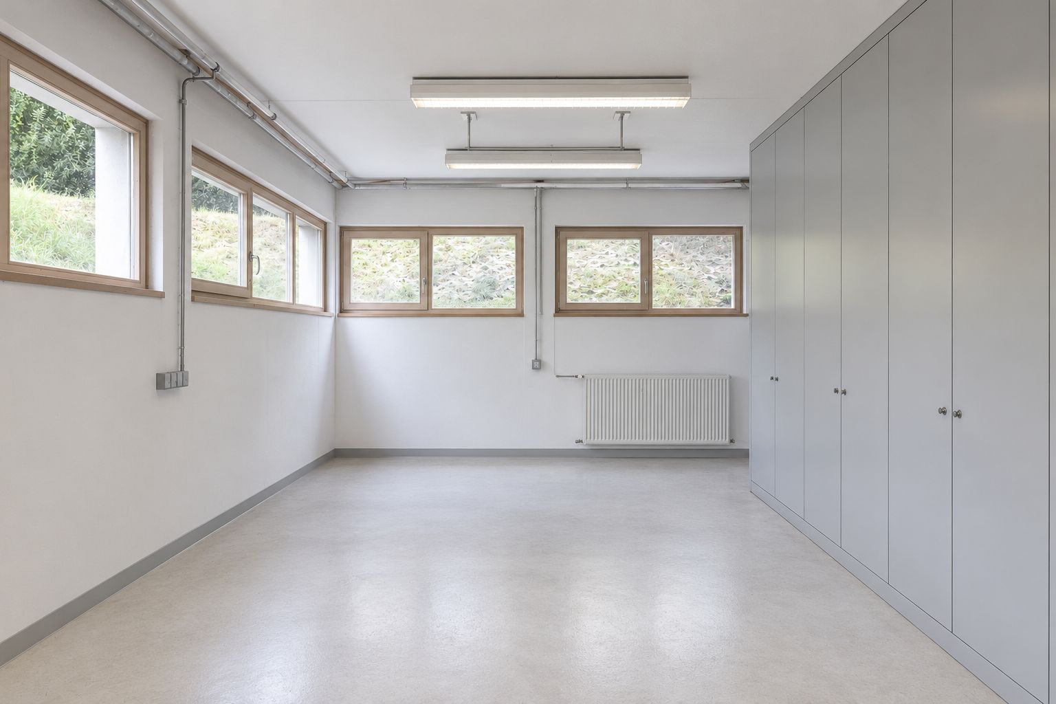Heller Raum mit mehreren Fenstern, grauem Boden und großen Einbauschränken an der rechten Wand.