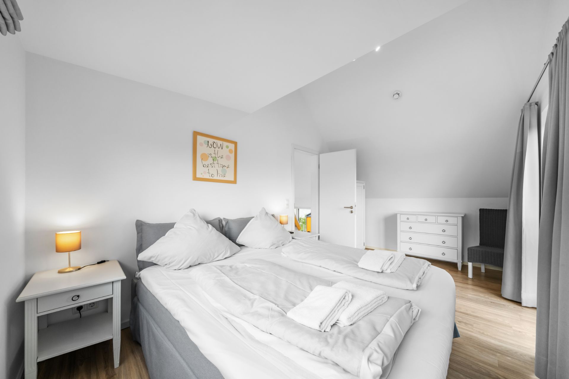 Helles Schlafzimmer mit Doppelbett, Nachttisch, Kommode und Stuhl auf Holzfußboden.