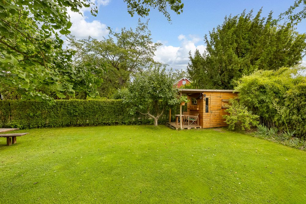 Garten mit Holzhaus, grünem Rasen, Bäumen und dichter Hecke im Hintergrund.