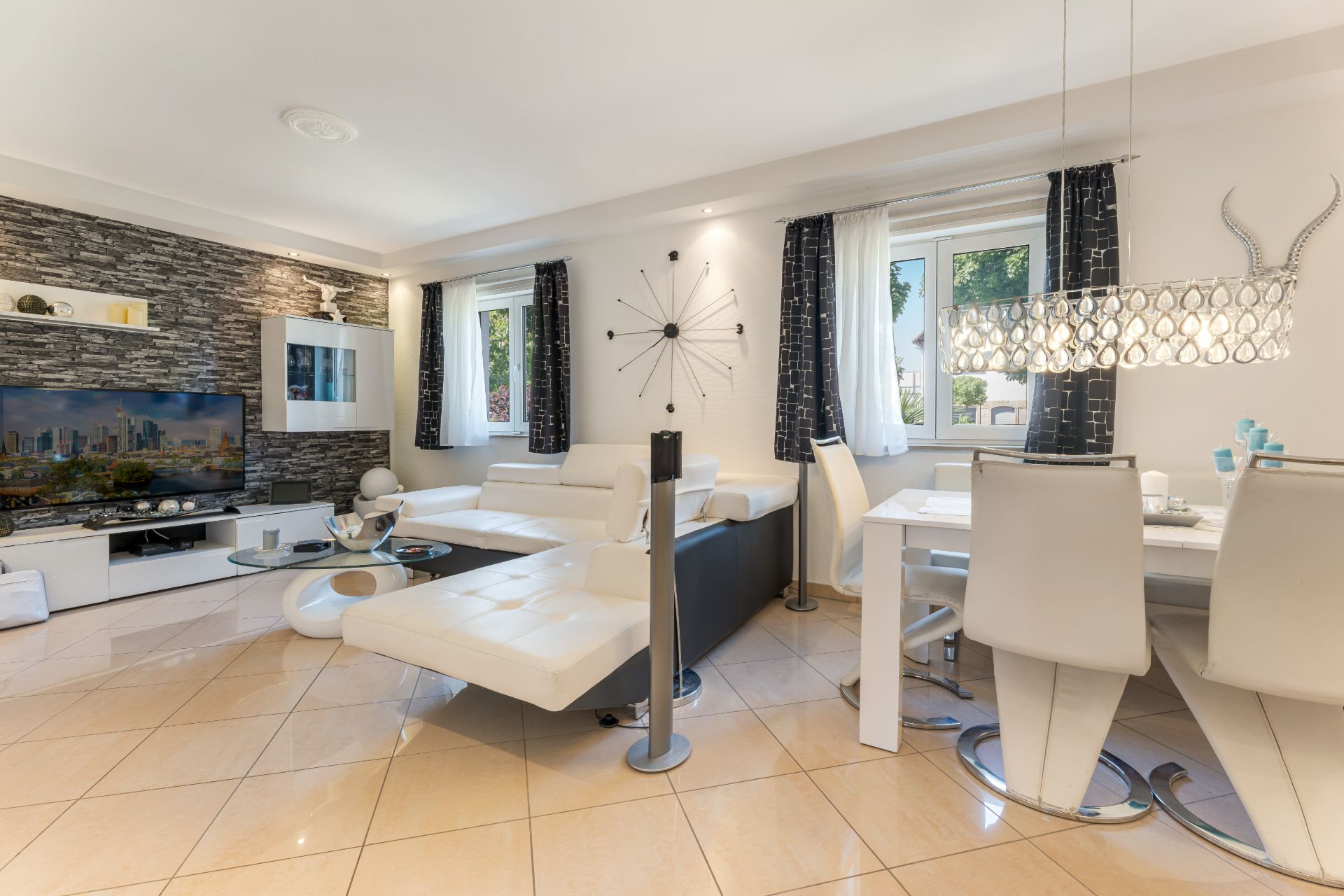 Helles Wohnzimmer mit weißem Sofa, Esstisch, Fliesenboden und großen Fenstern.