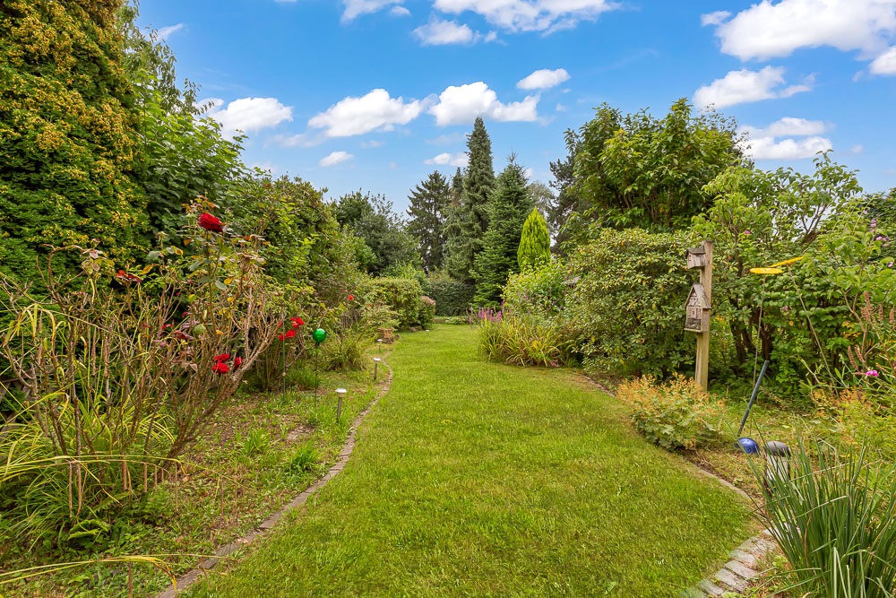 Grüner Garten mit Rasenweg, Blumen und Büschen unter blauem Himmel mit Wolken.
