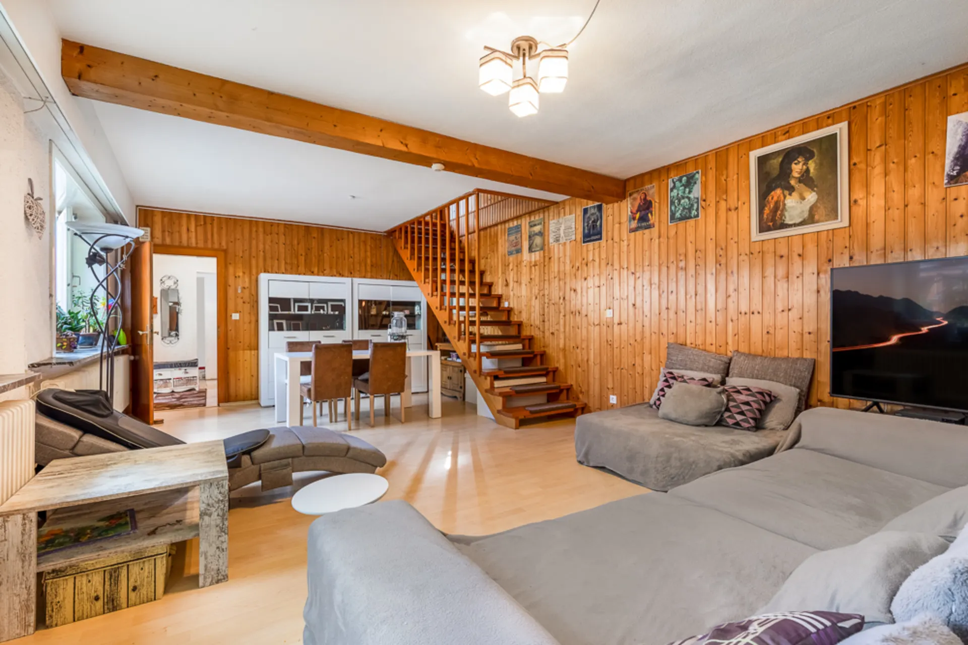 Wohnzimmer mit Holzvertäfelung, grauem Sofa, Esstisch, Fernseher und Treppe nach oben.