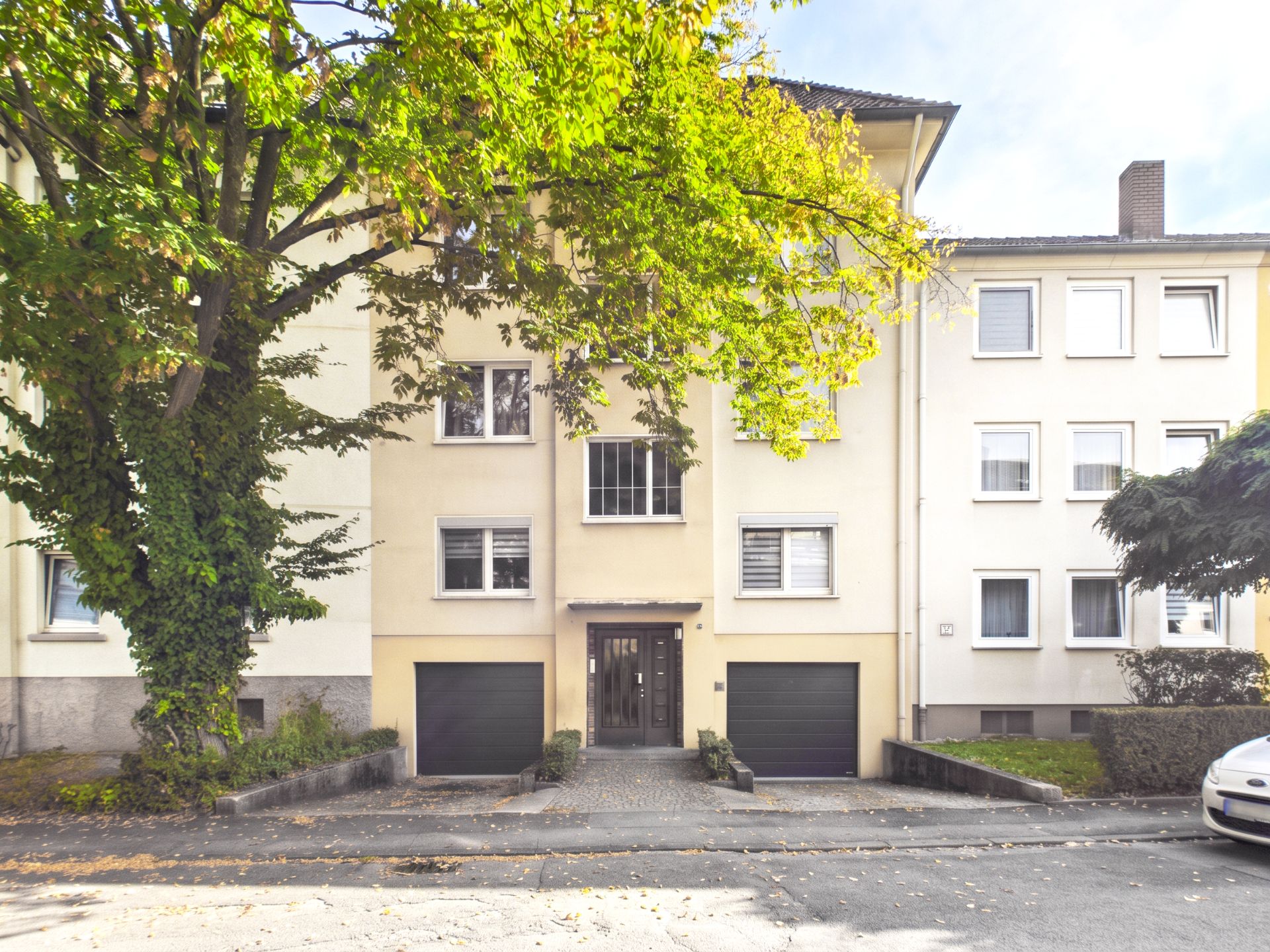 Mehrfamilienhaus mit zwei Garagen, großem Baum davor und mehreren Fenstern.