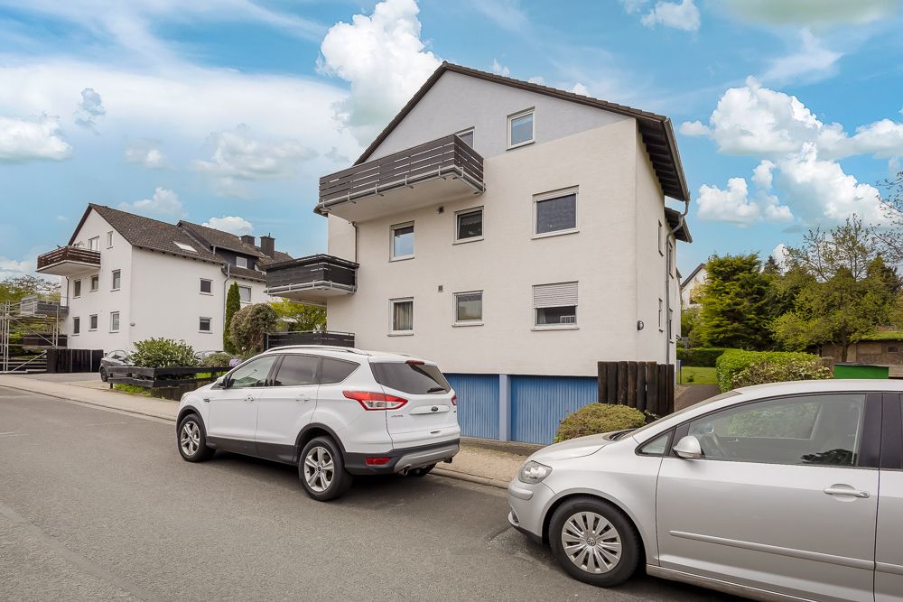 Weißes Wohnhaus mit Balkonen, davor zwei geparkte Autos auf einer ruhigen Straße.
