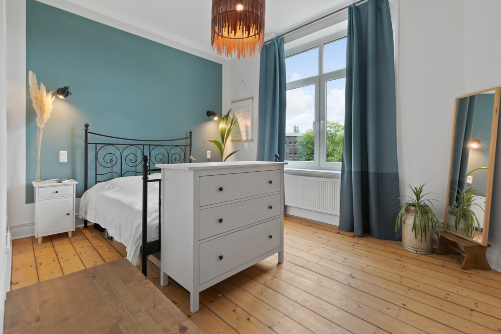 Helles Schlafzimmer mit Holzboden, Metallbett, Kommode, Spiegel und großen Fenstern.