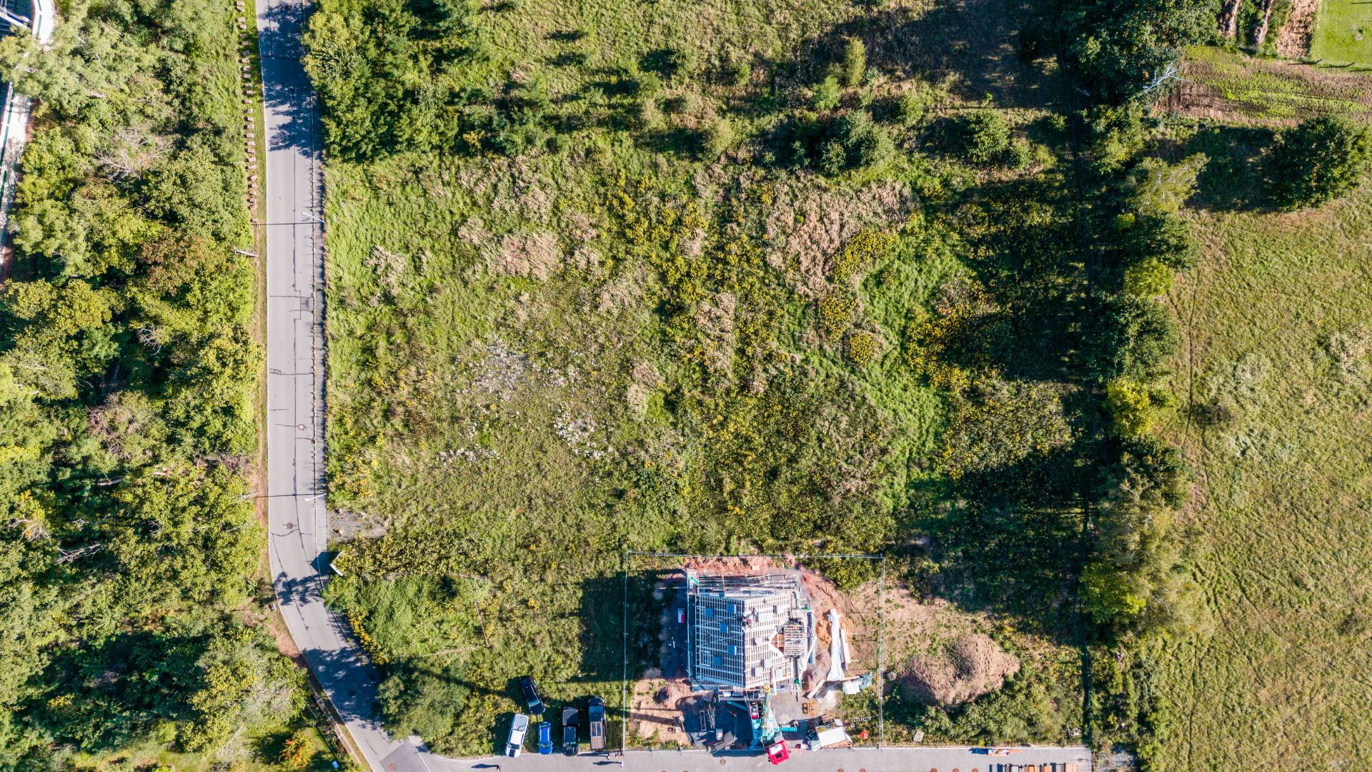 Luftaufnahme eines Grundstücks mit teilfertigem Haus, umgeben von Grünfläche und angrenzender Straße.