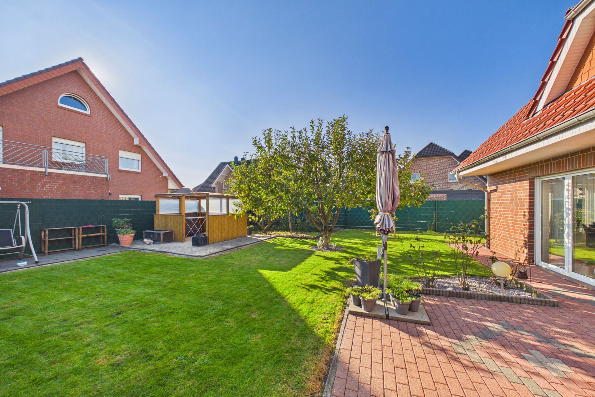 Grüner Garten mit Baum, Terrasse, Sonnenschirm und Gartenhaus an einem Wohnhaus.