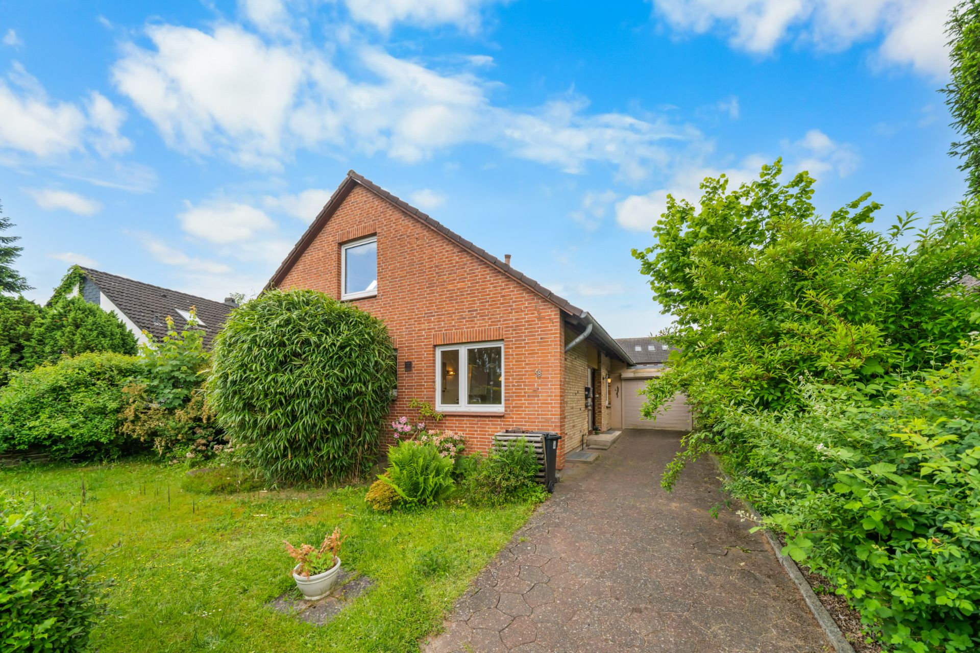 Backsteinhaus mit Garten, Einfahrt und Garage, umgeben von viel Grün unter blauem Himmel.