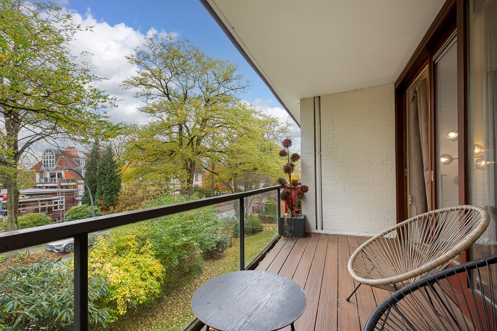 Balkon mit Holzboden, zwei runden Stühlen, Tisch und Blick auf grüne Bäume.