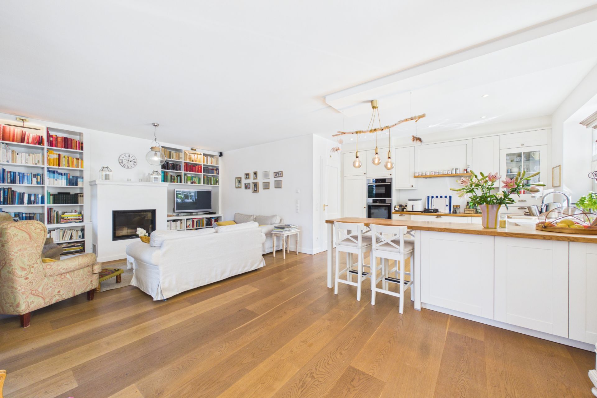 Helles Wohnzimmer mit offener Küche, Holzboden, Bücherregalen, Sofa und Kücheninsel mit Stühlen.