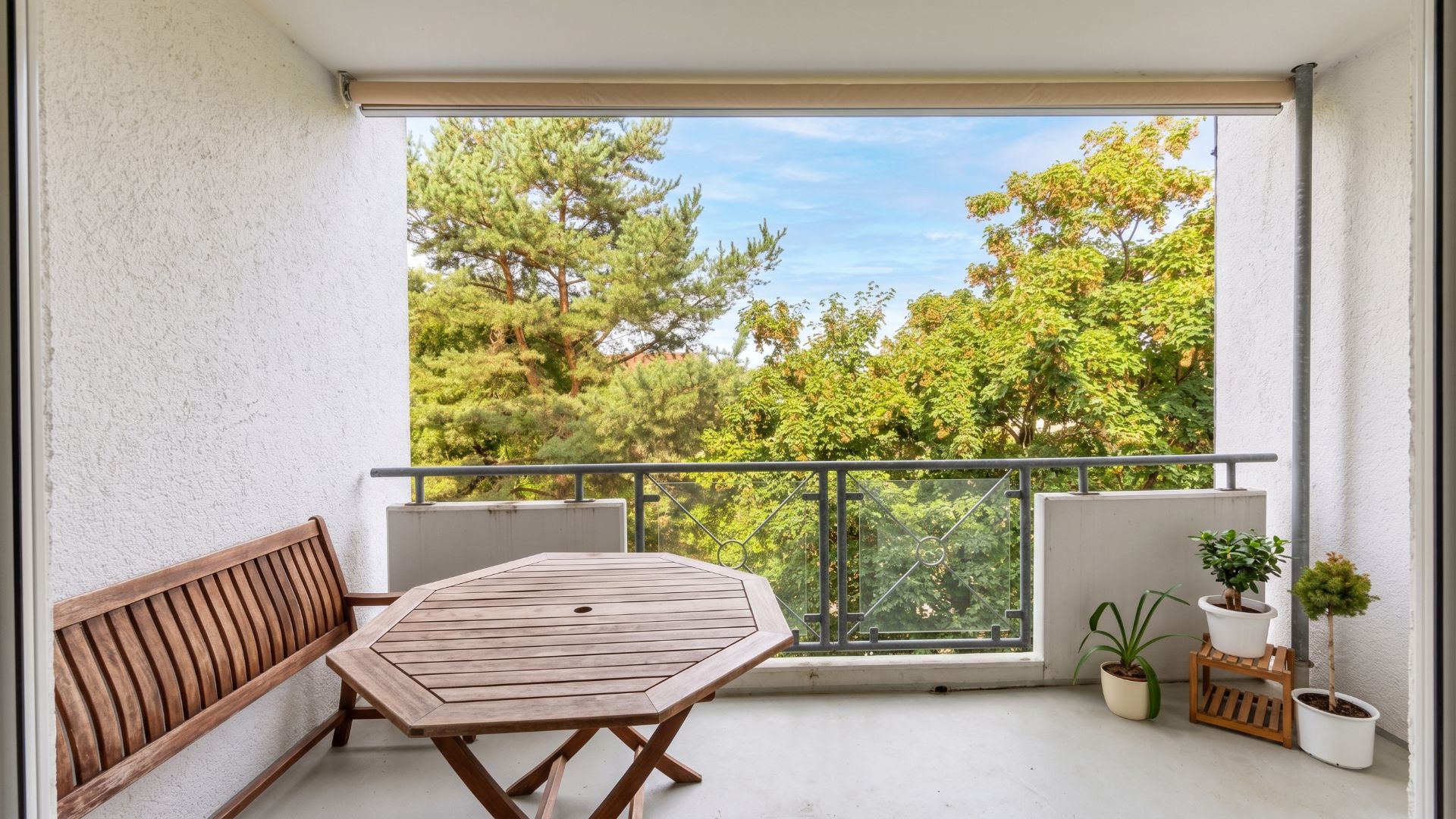 Überdachter Balkon mit Holztisch, Bank und mehreren Topfpflanzen, Blick ins Grüne.