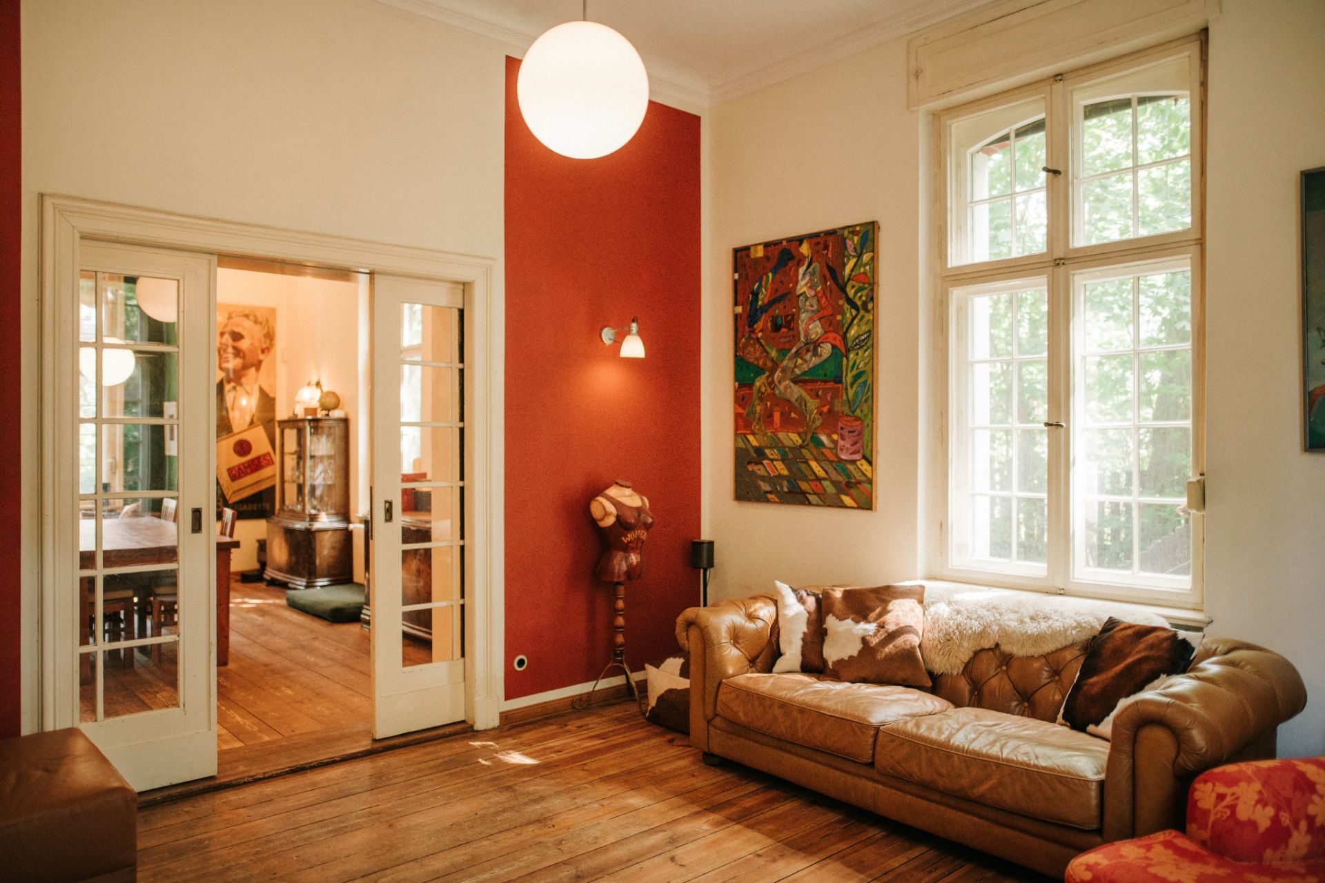 Helles Wohnzimmer mit Ledersofa, großem Fenster und bunter Wandkunst, Holzboden und Glastüren.