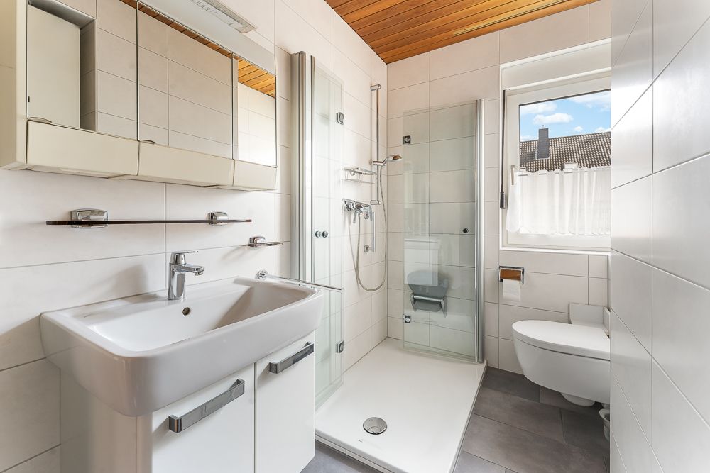 Modernes Badezimmer mit Waschbecken, Spiegel, ebenerdiger Dusche und WC, Tageslicht durch ein Fenster.