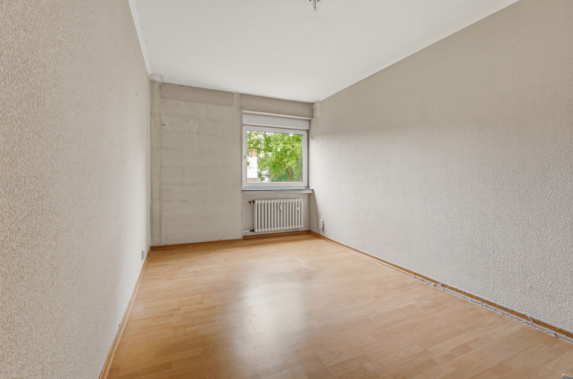 Leerer Raum mit Holzboden, weißer Wand und Fenster mit Blick ins Grüne.