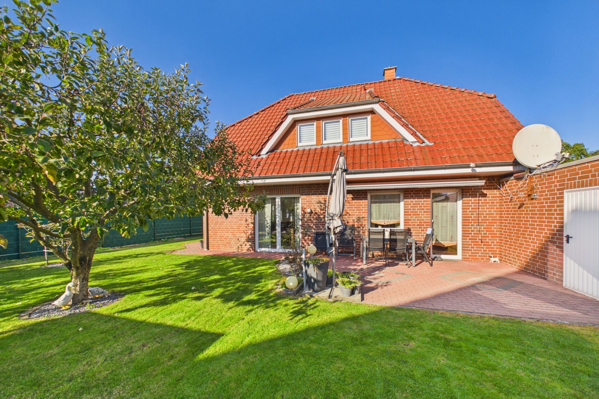 Backsteinwohnhaus mit rotem Dach, Terrasse, Gartenmöbeln und großem grünen Garten.
