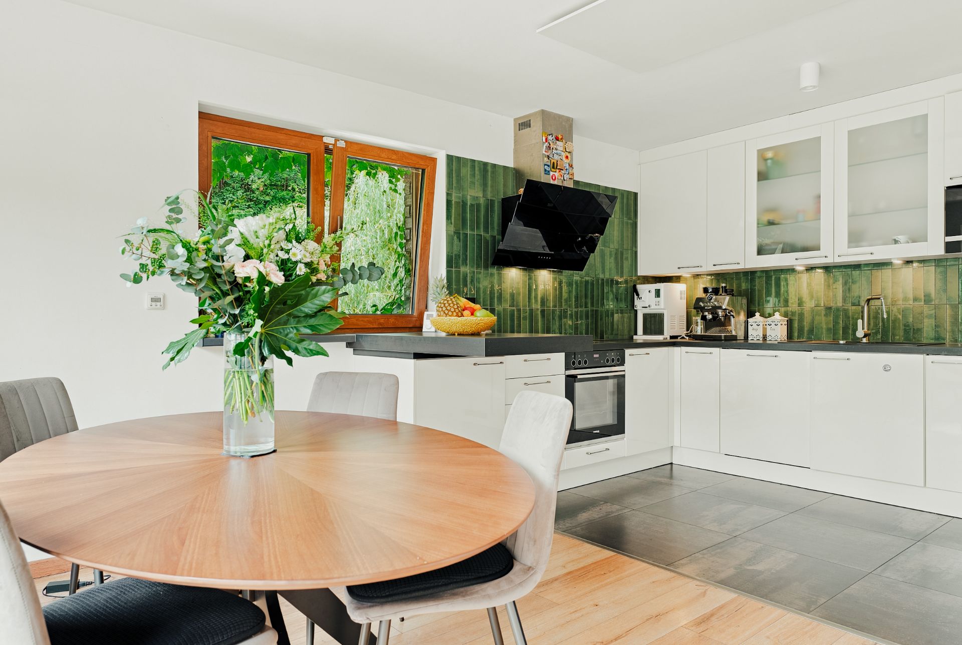 Helle Wohnküche mit rundem Tisch, Blumenstrauß und moderner Einbauküche mit grüner Fliesenwand.