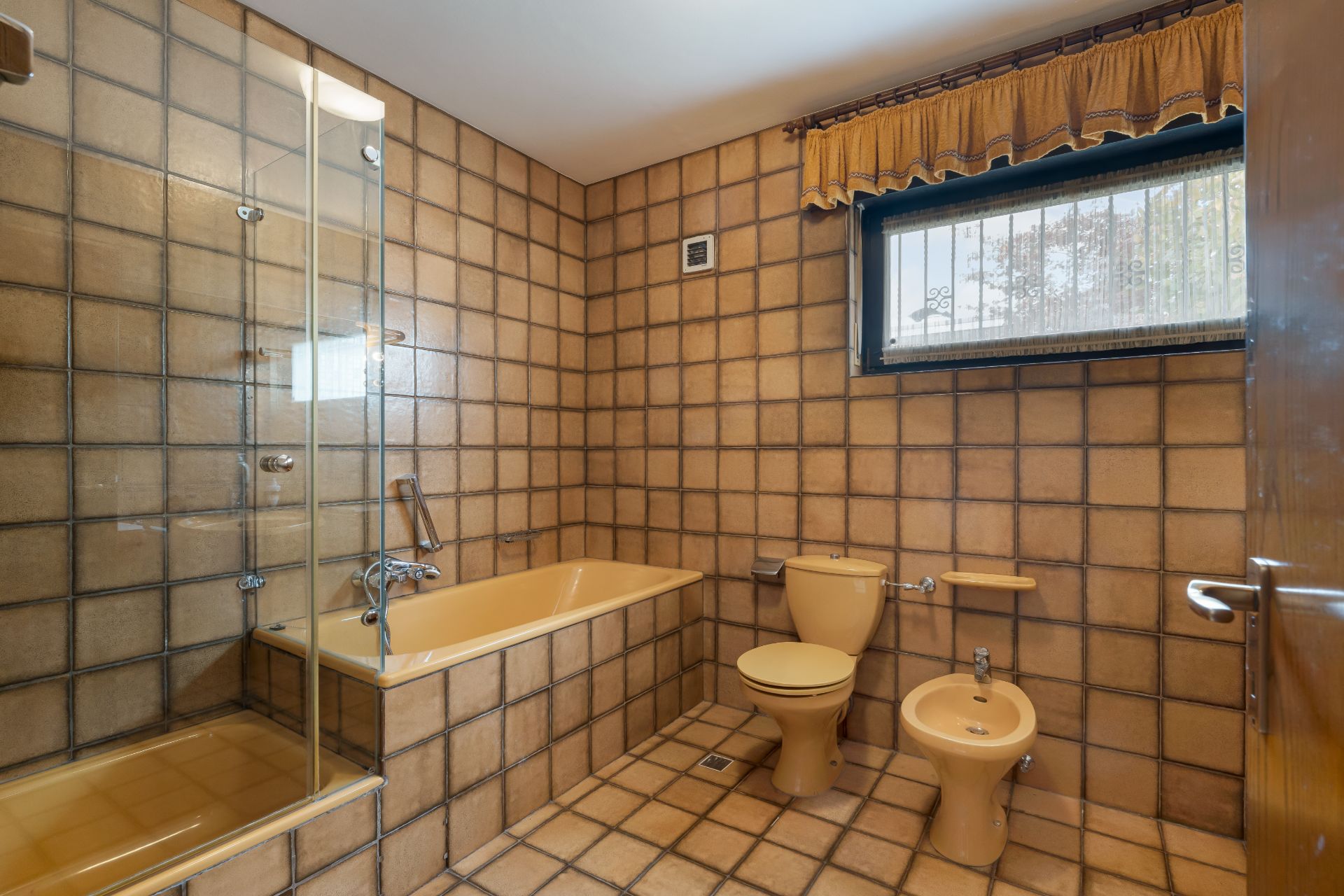 Badezimmer mit Badewanne, Toilette, Bidet und Dusche, braune Fliesen, Fenster mit Vorhang.