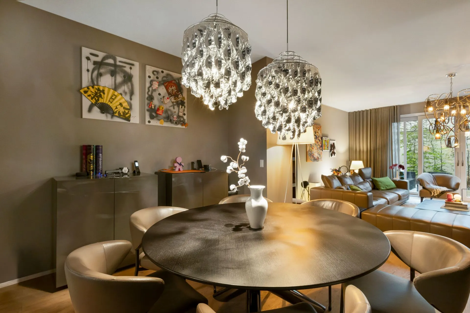 Modernes Wohnzimmer mit rundem Esstisch, Lederstühlen und funkelnden Kronleuchtern.