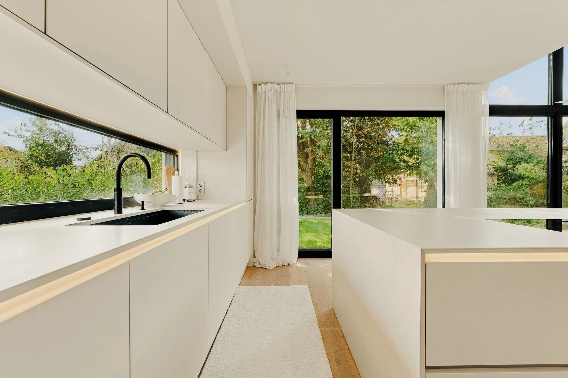 Helle, moderne Küche mit weißen Schränken, großer Insel und Blick auf den Garten durch große Fenster.