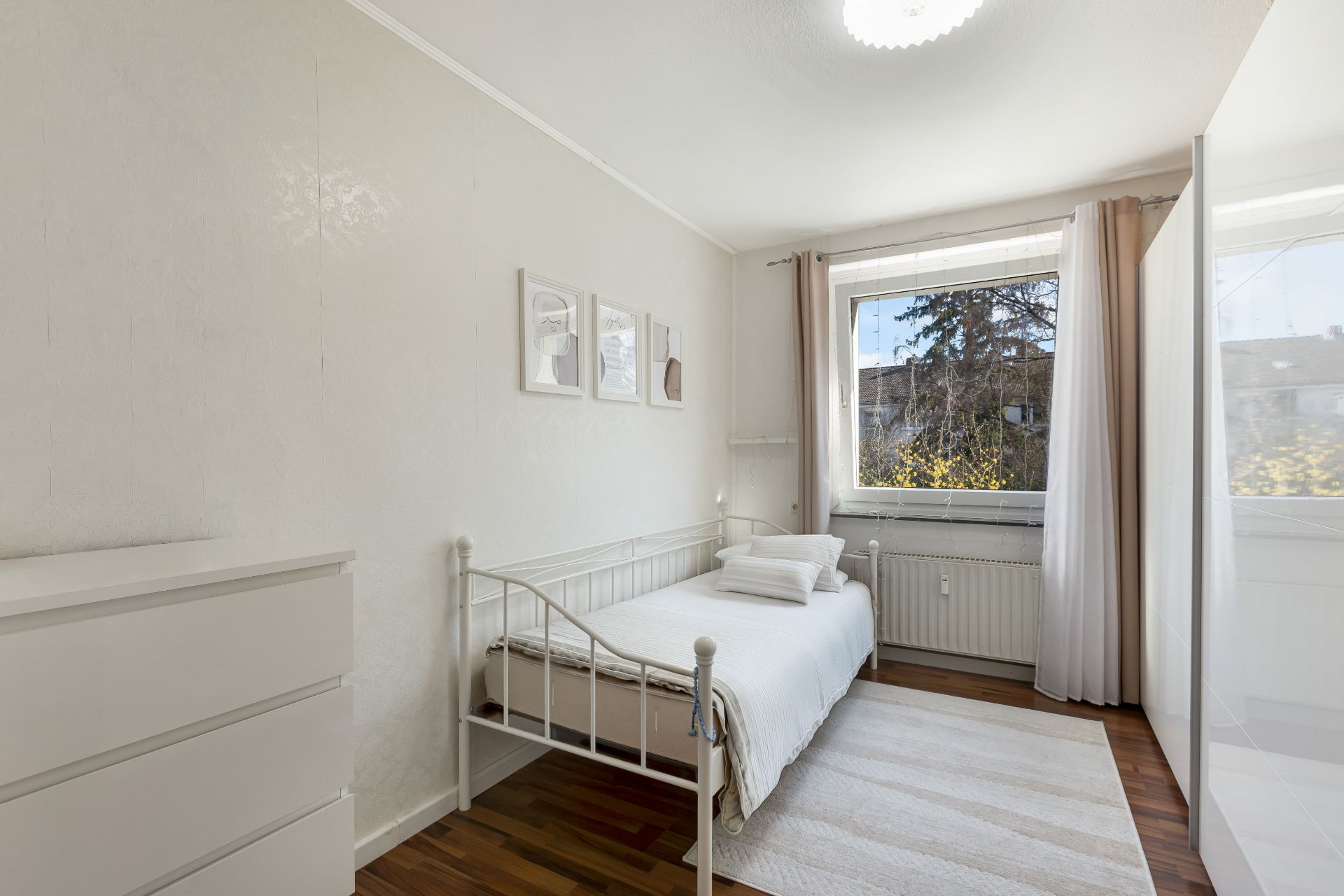 Helles Schlafzimmer mit Einzelbett, weißer Kommode, Teppich und großem Fenster mit Vorhängen.