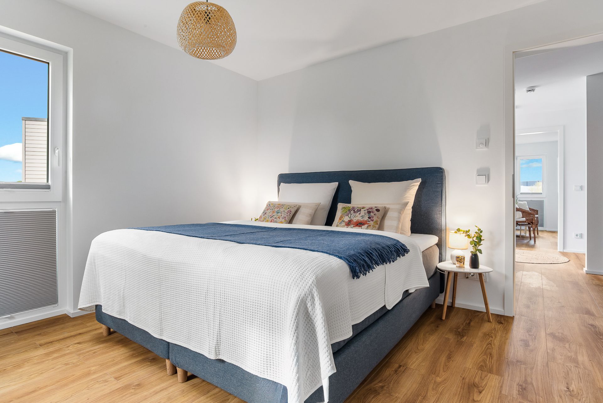 Helles Schlafzimmer mit Doppelbett, Holzfußboden, Nachttisch und großem Fenster.