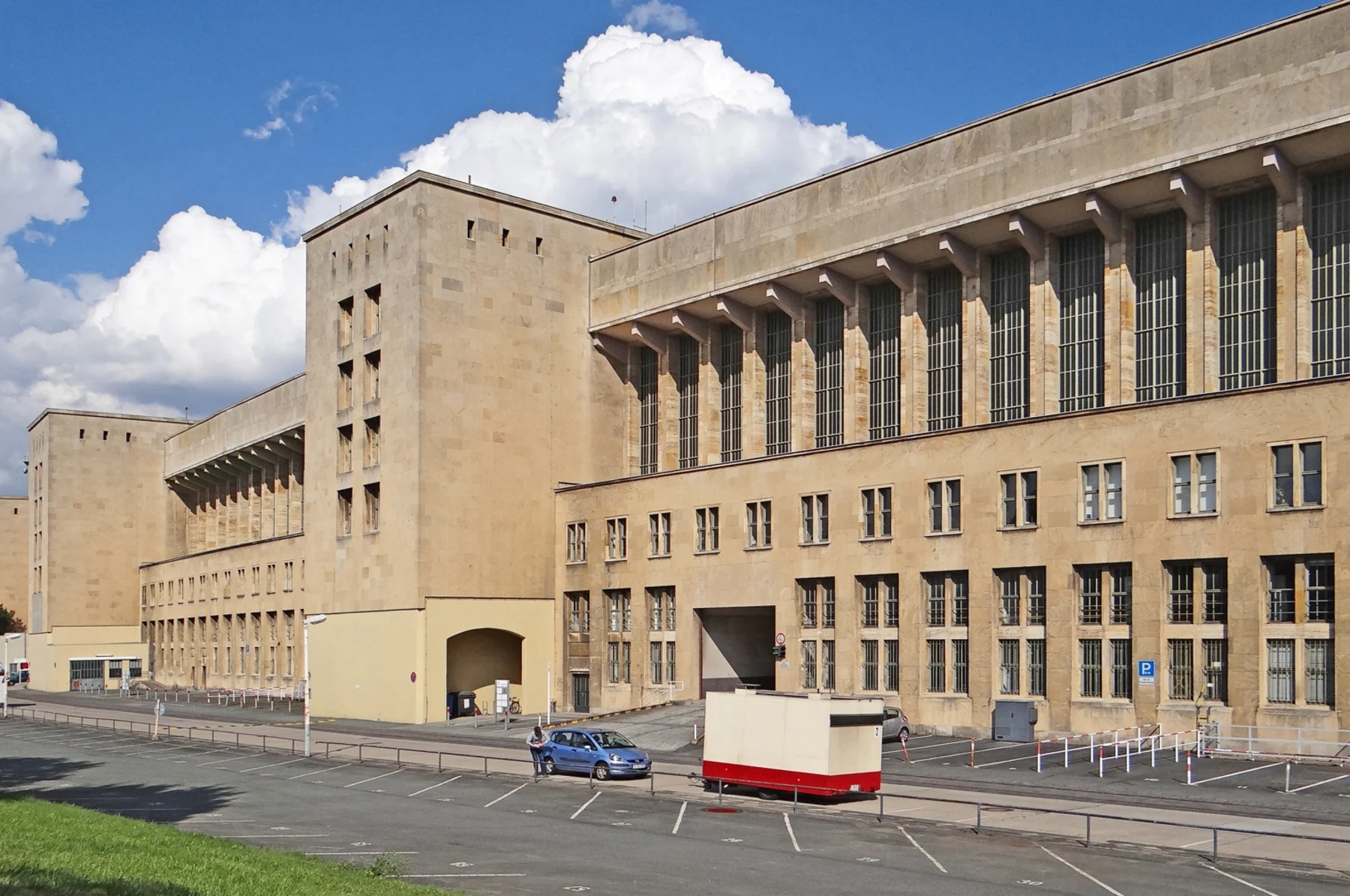 Großes, mehrstöckiges Gebäude aus hellem Stein mit vielen Fenstern und leerem Parkplatz davor.