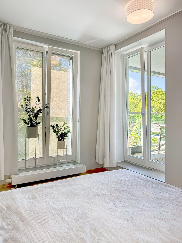 Helles Schlafzimmer mit zwei Fenstern, weißen Vorhängen und zwei Pflanzen auf Fensterbänken.