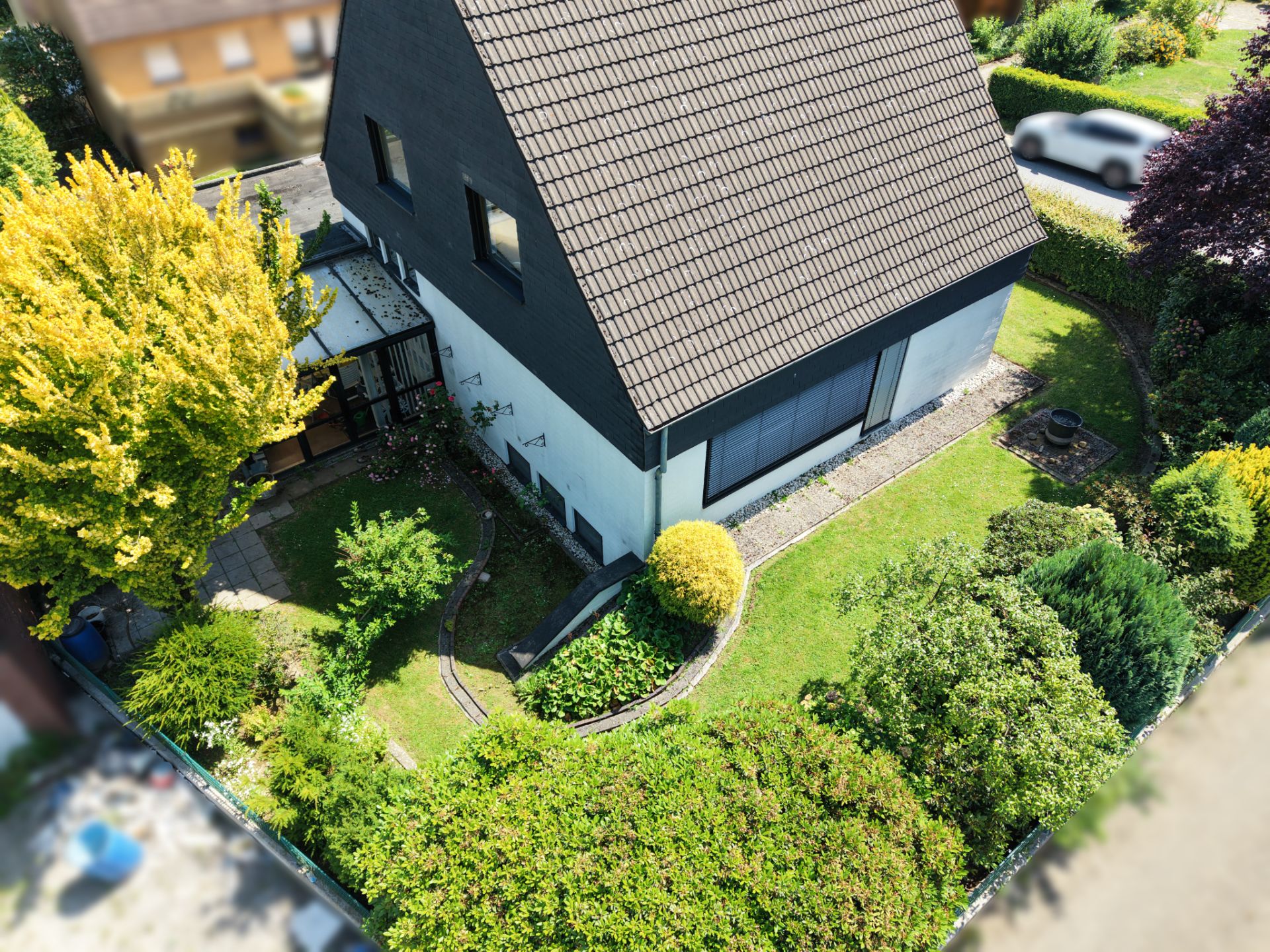 Luftaufnahme eines Einfamilienhauses mit Garten, Wegen und umgebenden Büschen und Bäumen.