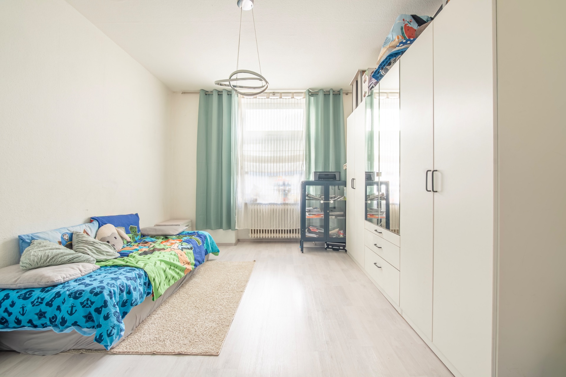 Helles Kinderzimmer mit Bett, Teppich, Kleiderschrank und Fenster mit grünen Vorhängen.