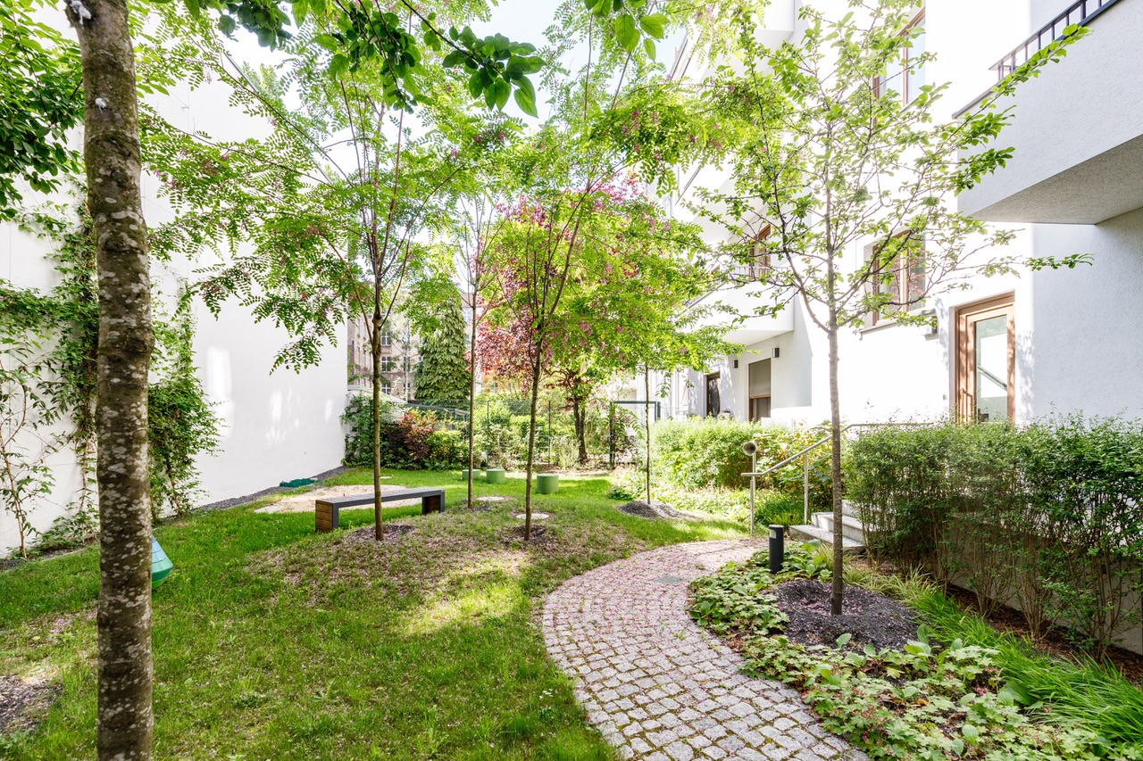 Gepflegter Garten mit Bäumen, Rasen, Weg aus Pflastersteinen und einer Bank bei Wohnhäusern.