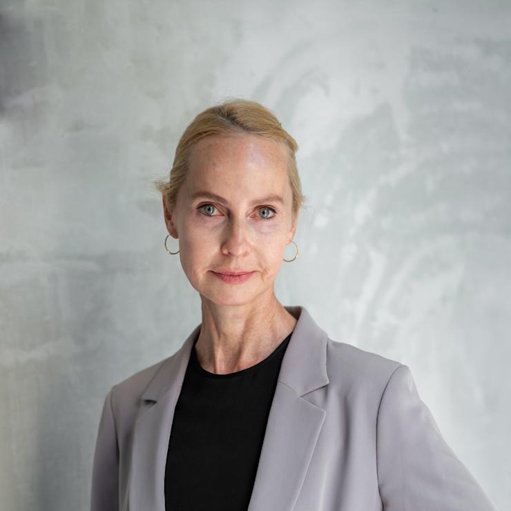 Frau mit blondem Haar, grauem Blazer und schwarzem Shirt steht vor einer grauen Wand.