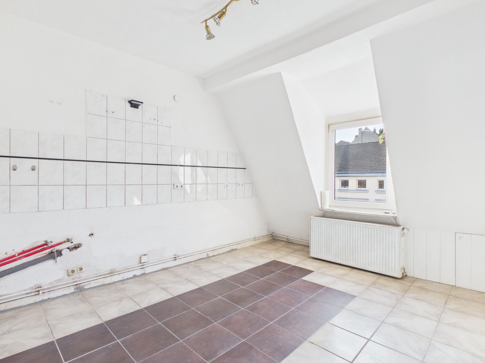 Leerer Raum mit weißen Fliesen, einem Fenster und Heizkörper, Anschlüsse an der Wand sichtbar.