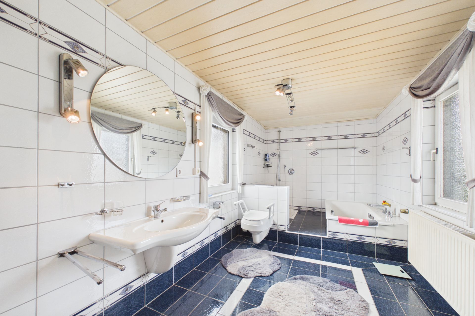 Helles Badezimmer mit Waschbecken, rundem Spiegel, Toilette, Badewanne und zwei Fenstern.