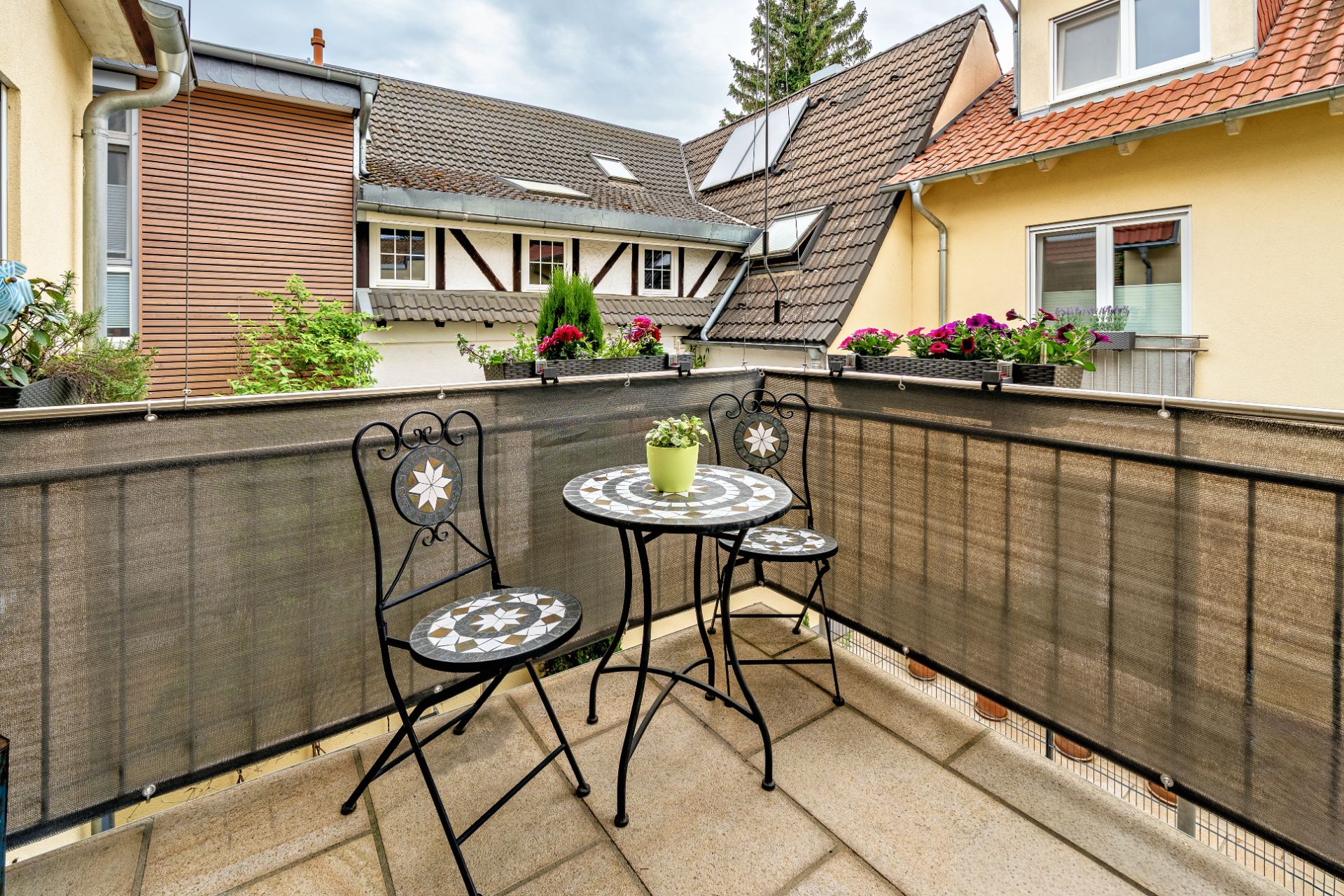 Kleiner Balkon mit zwei Stühlen, einem runden Tisch und Blumenkästen am Geländer.